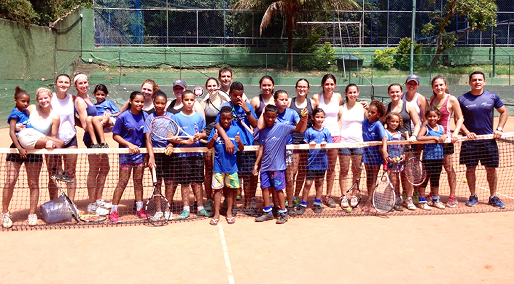 The Emory women’s tennis team pauses practice for a group photo with children in Brazil. The team traveled to Rio de Janeiro over the summer to improve their own tennis skills and volunteer their time to help children learn the sport. | Courtesy of Emory Athletics.